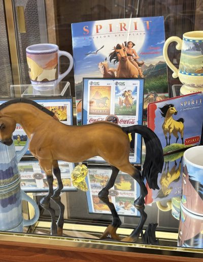 Collection of "Spirit: Stallion of the Cimarron" merchandise including a horse model, mugs, a poster, and coin set on a glass shelf.