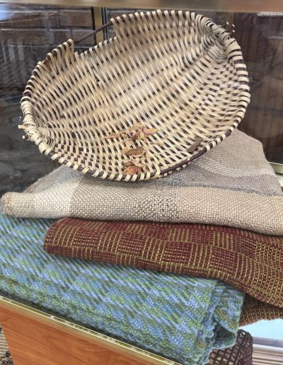 Woven oval basket with dried plant material displayed on layered, colorful textiles in a glass case.
