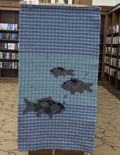 Blue woven wall hanging with three fish appliqués displayed in front of library bookshelves.